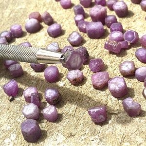 Könnte beinhalten: Eine Sammlung kleiner, facettierter, violetter Edelsteine, wahrscheinlich Rubine, die auf einer hellbraunen Oberfläche verstreut sind. Einige der Edelsteine sind in einer Reihe angeordnet, während andere zufällig verstreut sind. Ein kleines, silbernes Werkzeug ist auf dem Bild zu sehen.