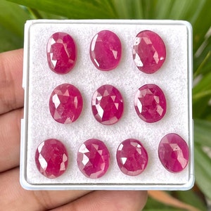 Puede incluir: Una colección de diez piedras preciosas de rubí facetadas en una caja blanca. Los rubíes son de forma ovalada y tienen un color rojo intenso.