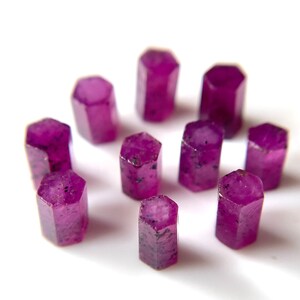 Może przedstawiać: Dziesięć fasetowanych rubinów, każdy o sześciokątnym kształcie, ułożonych na białym tle. Rubiny mają głęboki, bogaty purpurowy kolor i lekko prześwitują.