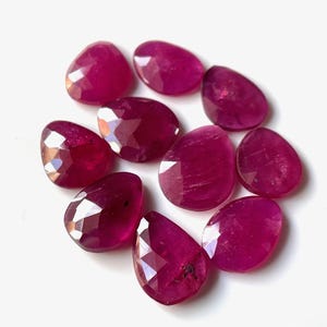 May include: A selection of faceted, teardrop-shaped ruby gemstones. The gems are a deep, rich red colour with a polished, reflective surface. The stones are arranged on a white background.