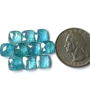 May include: A collection of ten square-cut, light blue gemstones next to a U.S. quarter. The gemstones have a faceted surface, reflecting light. The quarter is dated 1998.