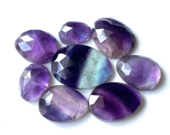 May include: A collection of eight faceted fluorite stones in shades of purple, green, and white. The stones are smooth and polished, with a variety of shapes and sizes.