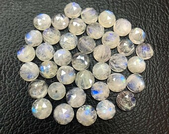 May include: A collection of faceted, round moonstone gemstones. The stones are mostly translucent with flashes of blue and white, and are arranged on a dark, textured surface. The gemstones are ready for jewelry making.