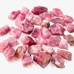 Puede incluir: Una colección de cristales de rubí rosa en bruto. Los cristales son de varios tamaños y formas, algunos tienen una apariencia más opaca que otros.