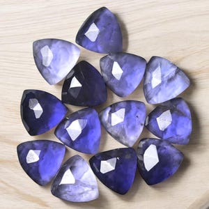 Op de afbeelding: Een verzameling van twaalf driehoekige gefacetteerde edelstenen in tinten paars en blauw. De stenen hebben een glinsterende, iriserende kwaliteit.