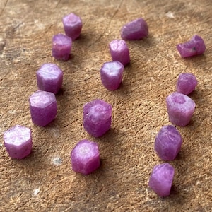 Puede incluir: Una colección de gemas de rubí en bruto, sin cortar. Las gemas son de un color rosa-rojizo vibrante y tienen forma hexagonal. Están dispersas sobre una superficie de madera rugosa, mostrando su estado natural y sin pulir. Ideales para joyería o coleccionismo.