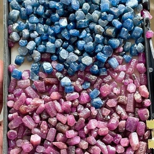 May include: A tray filled with raw gemstones. The top half is filled with blue gemstones, ranging from light to dark shades. The bottom half is filled with pink and red gemstones. The gemstones are rough-cut and have a natural, unpolished appearance.