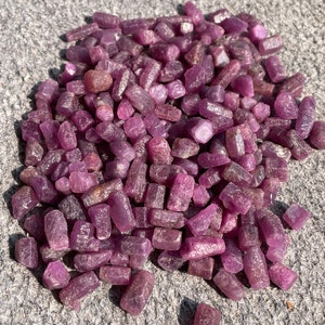 May include: A large pile of rough, pink ruby gemstones. The rubies are various sizes and shapes, with a natural, unpolished finish.