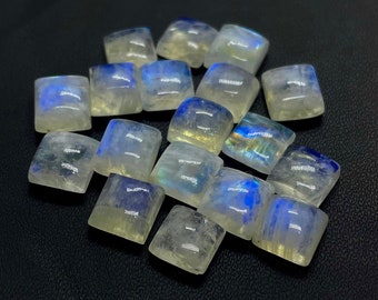 Könnte beinhalten: Eine Gruppe von 15 regenbogenfarbenen weißen Mondstein-Cabochons, jeder mit einer quadratischen Form und einer glatten, polierten Oberfläche. Die Steine haben einen dezenten blauen Schimmer und sind in einem lockeren Haufen angeordnet.