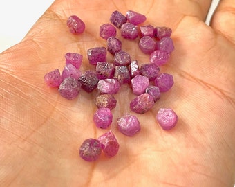 Può includere: Una collezione di piccoli cristalli di rubino rosa di varie forme e dimensioni. I cristalli sono sfaccettati e hanno un aspetto naturale e grezzo.