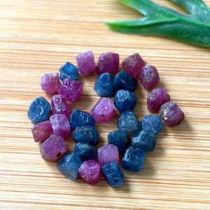 Può includere: Una collezione di piccole pietre preziose grezze e burattate in tonalità blu profondo e rosa vivace. Queste pietre individuali presentano forme irregolari e sfaccettate con una consistenza naturale non lucidata, sparse in una disposizione circolare.