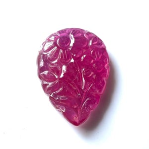 May include: A carved pink ruby gemstone pendant with a floral design. The pendant is shaped like a teardrop and has intricate details.