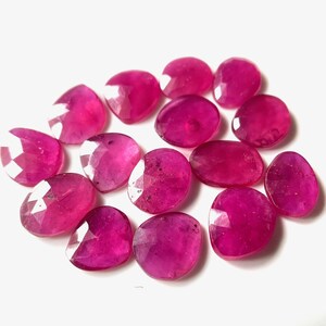 May include: A collection of 13 faceted ruby gemstones in a deep pink hue. The stones are oval shaped and have a smooth, polished finish.