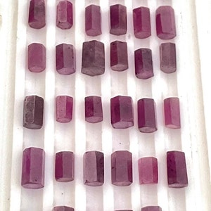 Op de afbeelding: Een verzameling van 30 gefacetteerde, paarse edelstenen gerangschikt in rijen op een wit oppervlak. De edelstenen zijn allemaal rechthoekig van vorm en hebben een gladde, gepolijste afwerking.