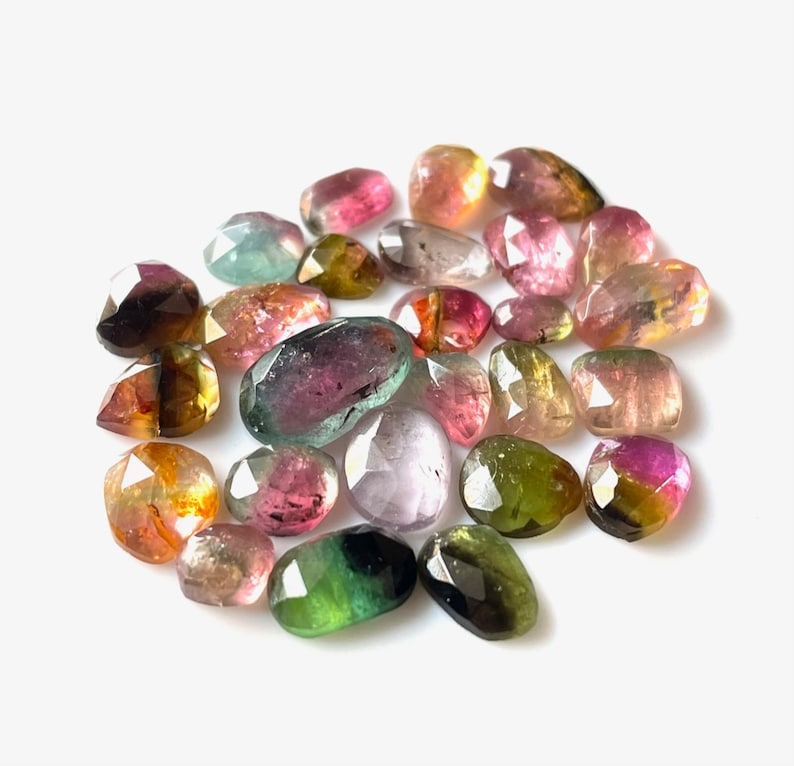May include: A collection of multi-colored, faceted, gemstone beads. The beads are various shades of pink, green, yellow, and black. The beads are oval shaped and have a smooth, polished finish.