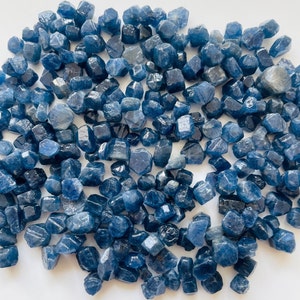 May include: A collection of small, faceted blue gemstones. The stones vary in shape and size, with a deep, rich blue color. The gemstones are scattered across a white surface, creating a visually appealing display. The overall effect is one of natural beauty and elegance.