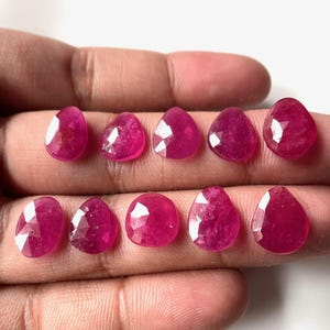 May include: Ten faceted ruby gemstone drops in a pear shape, displayed on a hand. The stones are a deep pink color.