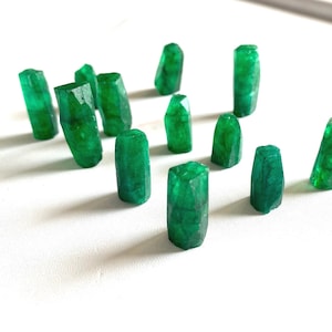 Puede incluir: Una colección de doce piedras preciosas de color verde esmeralda, talladas en forma rectangular, dispuestas sobre una superficie blanca.