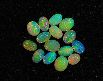 Puede incluir: Un conjunto de ópalos ovalados con colores vibrantes, incluyendo verde, azul y naranja, se encuentran sobre una superficie negra. Los ópalos muestran un efecto de juego de colores, reflejando la luz. Estas piedras preciosas son ideales para la creación de joyas.