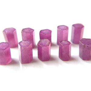 May include: Ten faceted pink ruby gemstones, each with a hexagonal shape, are arranged in a row on a white surface.