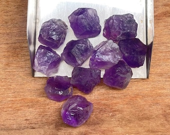 Peut inclure: Une collection de cristaux d'améthyste bruts, de tailles et de formes variées, présentés sur un plateau métallique. Les cristaux présentent une teinte violette foncée, avec des zones plus claires. Le fond est une surface en bois.
