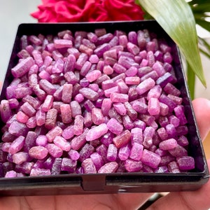Puede incluir: Una vista cercana de una pequeña caja negra llena de muchos pequeños cristales de rubí rosa sin cortar.