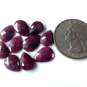 Peut inclure: Plusieurs pierres précieuses rouges en forme de poire et facettées. Une pièce de monnaie américaine est incluse pour la comparaison des tailles. Les pierres sont disposées sur une surface blanche, mettant en valeur leurs facettes polies.