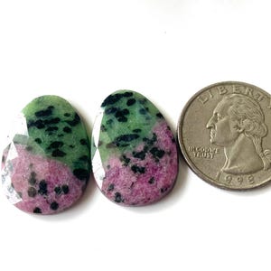 May include: Two oval-shaped gemstones with a faceted cut, featuring a mix of green, pink, and black colors. A U.S. quarter coin is next to the gemstones for size comparison. The gemstones are on a white surface.