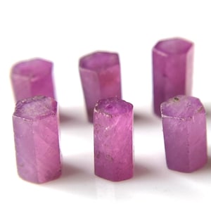 May include: Six faceted pink ruby gemstones, each with a hexagonal shape, are arranged in a circle on a white surface.