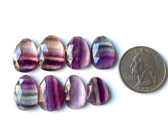 May include: Eight faceted, teardrop-shaped fluorite gemstones in shades of purple, lavender, and clear. A coin is next to the gemstones for size comparison. The gemstones have a polished, reflective surface.
