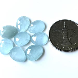 May include: A collection of eight light blue, faceted gemstones in an oval shape. A silver coin with Arabic script and a central design is also present. The gemstones are arranged on a white surface.