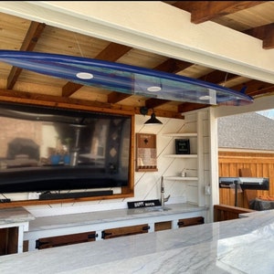 May include: An outdoor kitchen with a flat screen TV mounted on the wall, a white countertop, and a blue surfboard hanging from the ceiling. The kitchen has a sink, cabinets, and a grill.