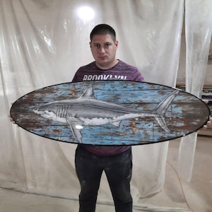 May include: Oval wooden wall art featuring a detailed illustration of a shark. The artwork has a distressed blue and brown background, giving it a rustic appearance. The shark is depicted in shades of gray and white.