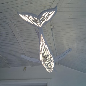 May include: A white whale-shaped ceiling light. The light has a cut-out design on the body and tail, creating a pattern when lit. The fixture is suspended from a white wooden ceiling.