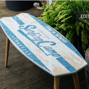 May include: A surfboard-shaped coffee table with a white and blue striped design. The table has the words "Surfing Camp" and "Living on the Beach, Los Angeles" printed on the top. It has four wooden legs.