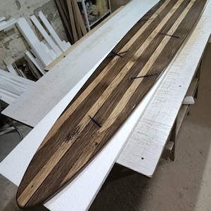 May include: A long, narrow wooden surfboard featuring a striped pattern of alternating dark brown and light tan wood grain. The board has a pointed nose and is constructed from multiple longitudinal wood strips joined together with visible dark cross-inlays.