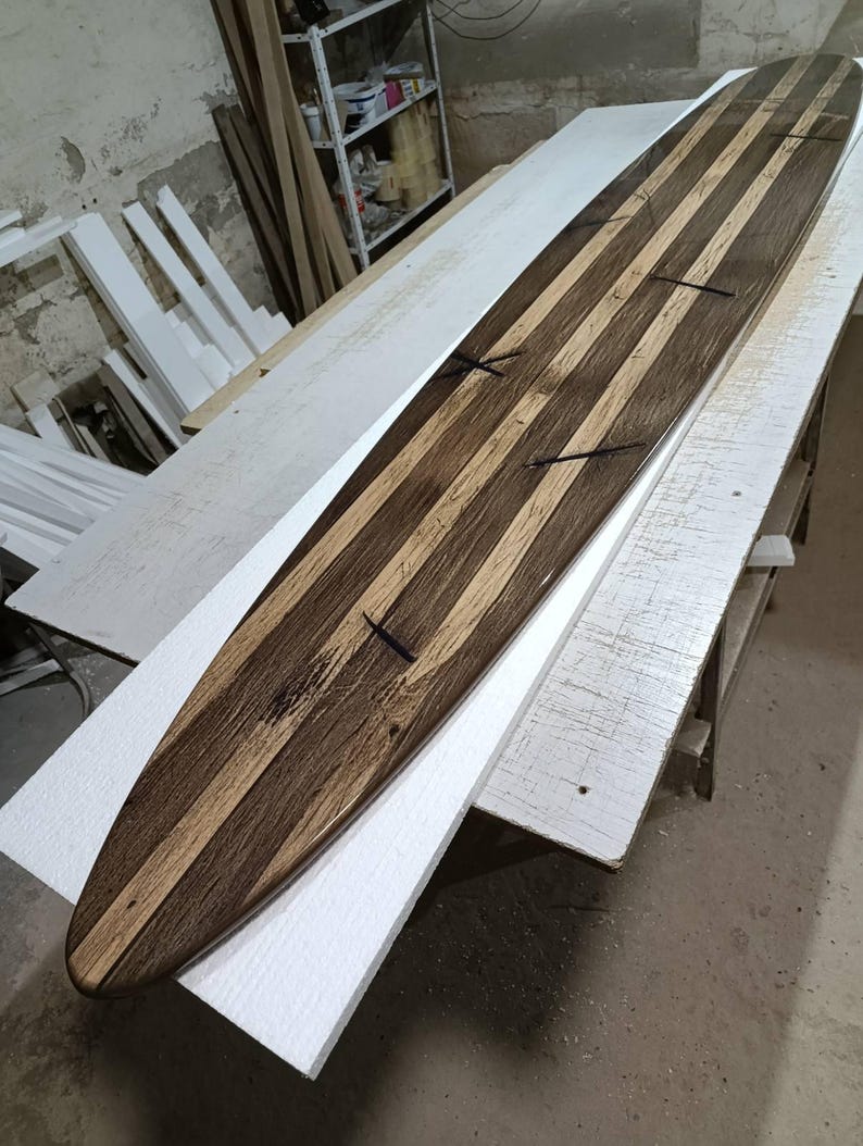 May include: A long, narrow wooden surfboard featuring a striped pattern of alternating dark brown and light tan wood grain. The board has a pointed nose and is constructed from multiple longitudinal wood strips joined together with visible dark cross-inlays.