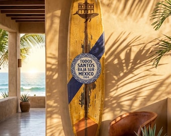May include: A long surfboard with a weathered yellow finish, a dark blue diagonal stripe, and a central circular emblem featuring the text "TODOS SANTOS BAJA SUR MEXICO 1964". The board displays a graphic of a building and a whale near the top.