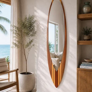 Puede incluir: Espejo grande con forma de tabla de surf con marco de madera. El marco presenta rayas verticales alternas de madera clara y oscura. El espejo está montado en una pared blanca.