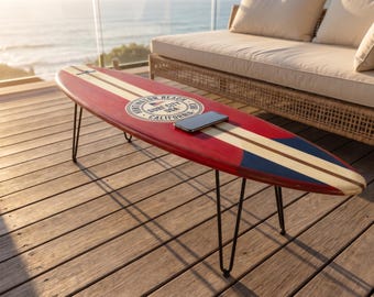 May include: A surfboard coffee table with black metal hairpin legs. The board is red with a central cream stripe and a blue tail section. A circular graphic reads Huntington Beach Surf City USA California. A black smartphone rests on the surface.