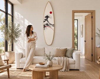 May include: A surfboard mounted on a wall featuring a graphic illustration of a large black and white ocean wave with a red circular sun and a thin red curved line accent against a cream-colored background with a wooden border.