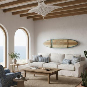 May include: A coastal-themed living room with a white sofa, striped pillows, and a wooden coffee table. A decorative manta ray light fixture hangs from the ceiling. A surfboard is mounted on the wall, and a patterned armchair sits in the corner.