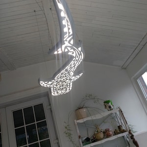 Könnte beinhalten: Eine hellgraue, abstrakte, dekorative Hängeskulptur mit komplizierten Ausschnittmustern. Die Skulptur ist von der Decke abgehängt und scheint aus einem hellfarbigen Material zu bestehen. Das Design wirft interessante Schatten.