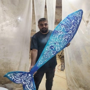 Puede incluir: Una gran escultura decorativa de cola de ballena. La cola está pintada en tonos de azul y blanco, con intrincados patrones y diseños. La escultura es sostenida por una persona en un taller.