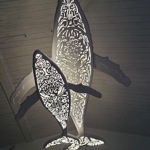 May include: Two illuminated whale sculptures hanging from a ceiling. The whales are white with intricate, cut-out patterns. The larger whale is positioned above a smaller one, both with fins and tail flukes. The light shines through the cut-outs.
