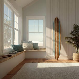 May include: A wooden surfboard with blue stripes leans against a white wall in a sunlit room. The room features a window seat with pillows, a large window, and a potted plant in a woven basket. The floor is hardwood and a light-colored rug.