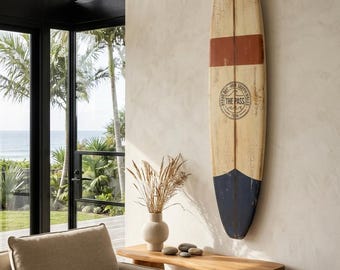 May include: A vintage-style surfboard mounted vertically on a wall, featuring a cream-colored body with a wide horizontal rust-orange stripe and a dark navy blue tail. The board displays a circular logo with the text THE PASS and wave graphics.