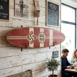 May include: A decorative wooden surfboard wall hanging featuring a distressed red and white striped finish, a carved Basque cross symbol, and a circular emblem with the text Cote des Basques 1991 Biarritz, mounted horizontally on black metal brackets.