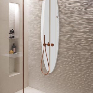 May include: A white surfboard-shaped mirror with vertical gold stripes and copper shower fixtures. The showerhead and handheld shower are copper-coloured. The mirror is mounted on a textured, wave-patterned wall in a modern bathroom.