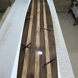 May include: A longboard deck featuring a striped pattern of alternating dark brown and light tan wood grain panels. The surface has a glossy, reflective finish and includes several dark, thin, cross-shaped inlay details embedded across the length.